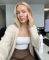 Woman applying cream to her face in an office setting with a bottle of 'Miller &;&'N'Boozy' product on the desk.